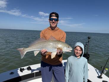 A proud angler and a young boy display a large Redfish aboard their boat, enjoying a sunny day on the water.