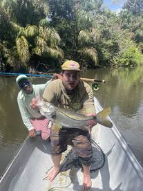 Angler proudly displaying a large Snook while fishing in a tropical river environment, accompanied by a fellow fisherman.
