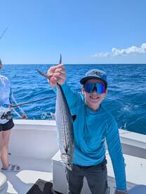 Young angler proudly holding a Spanish Mackerel while fishing on a boat in clear blue waters.