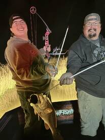 Two anglers proudly displaying a large bass caught during a nighttime bowfishing trip.