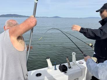 Two anglers actively fishing on a boat, with one reeling in a catch while the other assists. The serene water and distant hills create a picturesque fishing environment.