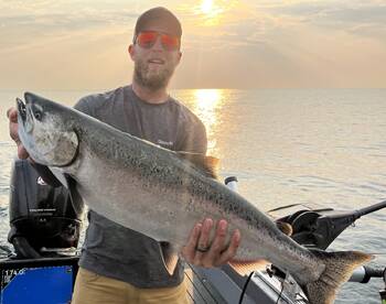 Angler proudly displaying a large Salmon against a stunning sunset backdrop while fishing on a boat.