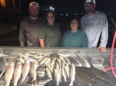 A group of four anglers proudly displaying their catch of various fish, including redfish and flounder, on a cleaning table at night.