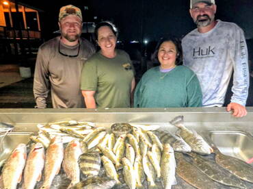 A group of four anglers proudly displaying a variety of fish, including Redfish and Speckled Trout, on a cleaning table after a successful night fishing trip.