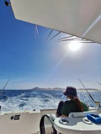 A young angler relaxing in the seating area of a fishing boat, enjoying a sunny day on the water with a scenic view of the coastline.