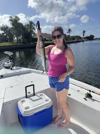 A smiling angler proudly displays her catch, a fish, while standing on a boat in a scenic waterway.