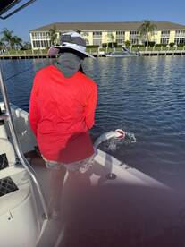 An angler in a red shirt is actively reeling in a fish from a boat in a scenic waterway, showcasing the excitement of fishing.