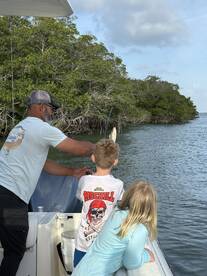 A young boy is reeling in a fish while an adult assists him on a boat surrounded by mangroves, showcasing a family fishing experience.