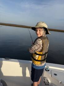 A young angler fishing from a boat in calm waters, wearing a life jacket and a wide-brimmed hat, enjoying a sunny day on the water.
