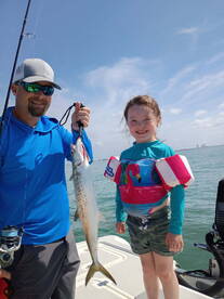 A young girl proudly poses with her father, showcasing a large Speckled Trout caught during a fun day of fishing on the water.