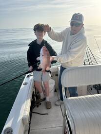 A young angler and an adult proudly displaying a large Red Snapper while fishing on a boat in calm waters.