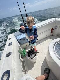 A young angler on a boat proudly displaying a caught fish, with a bucket full of additional catches nearby, showcasing a successful fishing trip.