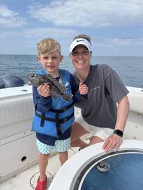 A young boy proudly holds a Black Sea Bass while fishing with his mother on a boat, showcasing a memorable family fishing experience.