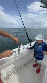 A young angler in a life jacket excitedly watches as an adult pulls a fish from the water during a fishing trip.