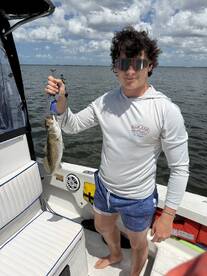Young angler proudly holding a Speckled Trout while fishing on a boat in a scenic waterway.