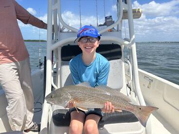 A young angler proudly displaying a large Redfish while seated on a boat, enjoying a sunny day on the water.