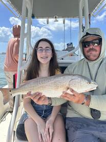 A young angler and an adult proudly displaying a large Redfish on a boat during a fishing trip.