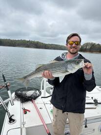Angler proudly displaying a large Striped Bass while fishing on a lake, showcasing a successful catch in a scenic environment.