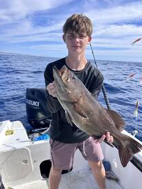 A young angler proudly displaying a large Grouper caught during a fishing trip on a boat in open waters.