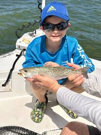 A young angler proudly displaying a Speckled Trout while fishing on a boat, showcasing a fun day on the water.
