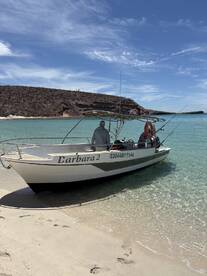 The 'Carbara 2' boat is anchored on a sandy beach, with two anglers preparing for a fishing trip in a scenic coastal environment.