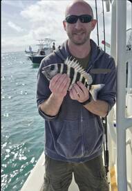 Angler proudly displaying a Sheepshead caught during a fishing trip, with boats visible in the background.