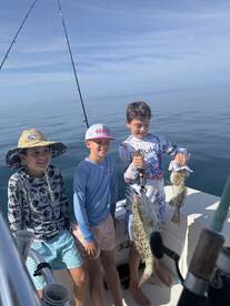 Three young anglers proudly displaying their catch of grouper while fishing on a boat in calm waters.