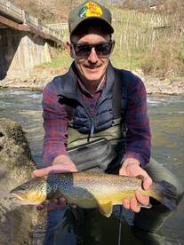 Angler proudly displaying a large Brown Trout while standing in a scenic river setting.