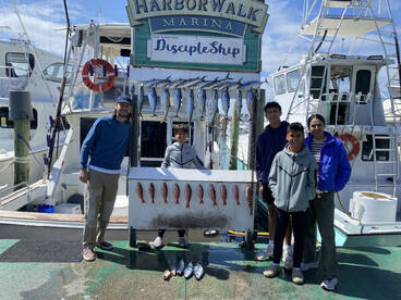 A group of young anglers proudly posing with their catch of various fish, including Snapper, displayed on a catch board at Harborwalk Marina.