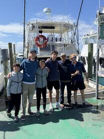 A group of five young anglers and a captain pose together in front of a charter boat at the marina, showcasing a fun day of fishing.