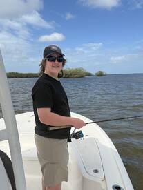 A young angler casting from the deck of a boat in a scenic fishing environment, surrounded by mangroves and open water.