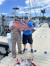 Two anglers proudly displaying a large Red Grouper at the marina after a successful fishing trip, with several smaller fish laid out on ice nearby.
