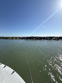 A fishing rod is positioned over the water, capturing the serene environment near a rocky jetty on a clear day.