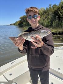 A young angler proudly displaying a Speckled Trout while fishing from a boat in a scenic coastal environment.