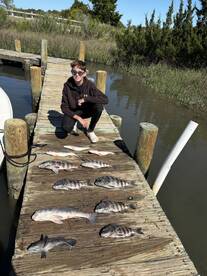 A young angler poses next to a variety of freshly caught fish on a dock, showcasing a successful day of fishing in a scenic environment.