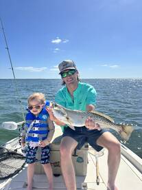 A young child and an adult proudly display a large Speckled Trout while fishing on a sunny day.