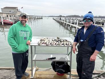 Two anglers proudly standing beside a catch of multiple fish displayed on a cleaning table at the dock, showcasing their successful fishing trip.