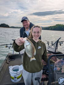 A young angler proudly displaying a Crappie while fishing with a family member on a scenic lake.