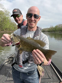 Angler proudly displaying a large Largemouth Bass while fishing on a river, with a fellow fisherman in the background.