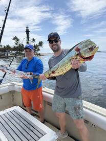 Two anglers proudly displaying their catch of a large Mahi Mahi and a Barracuda while fishing near a lighthouse.