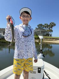 A young angler proudly displaying a caught fish while standing on a boat in a scenic waterway.