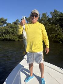 An angler proudly displaying a large Snook while standing on the bow of a boat in a mangrove setting.