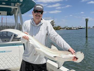 Angler proudly displaying a large shark caught during a fishing trip, with a scenic waterfront in the background.