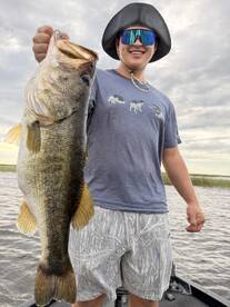 A young angler proudly displaying a large Largemouth Bass while fishing from a boat in a scenic freshwater environment.