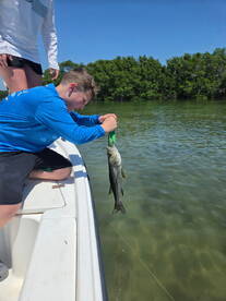 A young angler is reeling in a Snook while fishing from a boat in a scenic coastal environment.