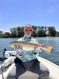 Angler proudly displaying a Redfish while fishing on a sunny day, surrounded by lush greenery along the water.