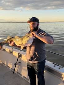 An angler proudly displaying a large Striped Bass while fishing on a boat during sunset.