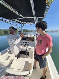 Two anglers discussing fishing techniques inside a boat, with a calm water backdrop and clear skies.