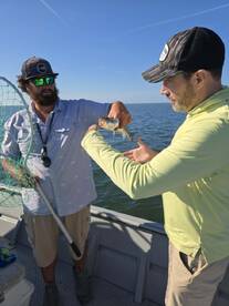 Two anglers on a boat, one proudly displaying a caught fish while the other assists, showcasing a successful fishing trip.