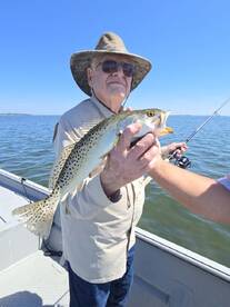 An elderly angler proudly displaying a Speckled Trout while fishing on a sunny day in a bay.